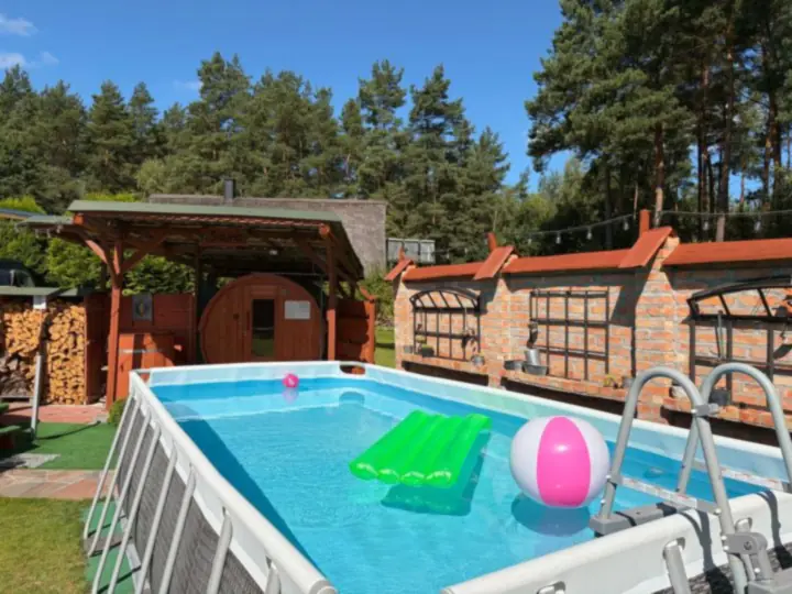 Prostokątny basen stelażowy z drabinką, materacem i piłką, w tle sauna i wiata