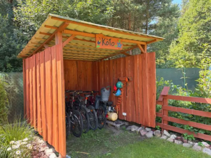 Rowery pod drewnianą wiatą z malowanym szyldem, obok płot i zieleń, słoneczny dzień