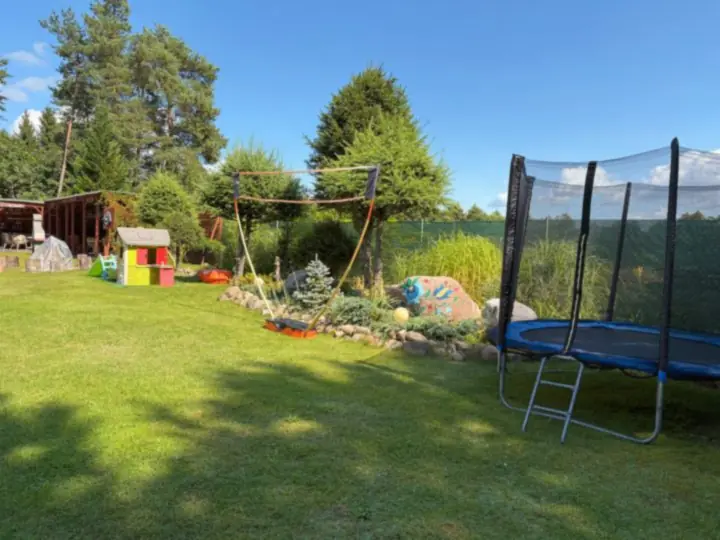 Trampolina z siatką na dużym trawniku, w tle domek do zabawy